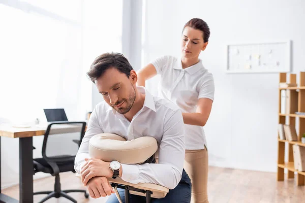 Massaggiatrice donna massaggiatrice schiena di uomo d'affari seduto su sedia da massaggio in ufficio su sfondo sfocato — Foto stock