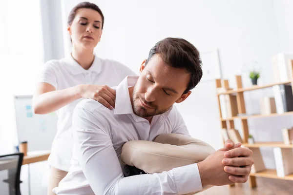 Massaggiatrice massaggiando spalle di uomo d'affari sorridente con gli occhi chiusi in ufficio su sfondo sfocato — Foto stock