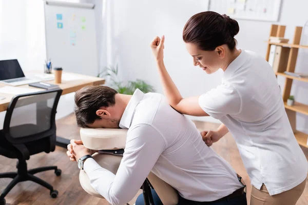 Masajista sonriente haciendo masaje de espalda con codo para hombre de negocios en oficina sobre fondo borroso - foto de stock