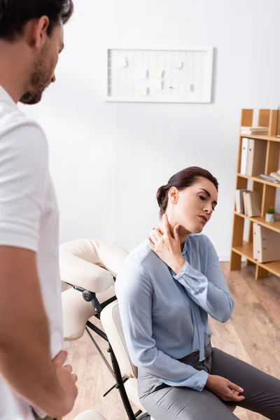 Masseur com a mão no quadril olhando para mulher de negócios com dor no pescoço sentado na cadeira de massagem no escritório em primeiro plano borrado — Fotografia de Stock