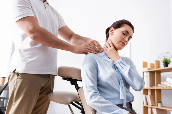 Masajista terapeuta masajeando hombro doloroso de mujer de negocios sentada en silla de masaje en la oficina sobre fondo borroso - foto de stock