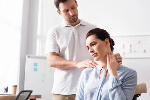 Massagetherapeutin massiert schmerzhaften Hals einer Geschäftsfrau mit verschwommenem Büro auf Hintergrund — Stockfoto