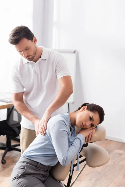 Massaggiatrice massaggiatrice massaggiando schiena di donna d'affari seduta su sedia da massaggio in ufficio su sfondo sfocato — Foto stock