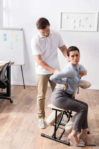 Masajista masajeando dolorosa espalda de mujer con la mano en la cadera, sentado en silla de masaje en la oficina - foto de stock