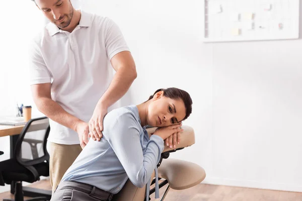Massagista massagista sorridente massageando costas de empresária sentada na cadeira de massagem no escritório no fundo borrado — Fotografia de Stock