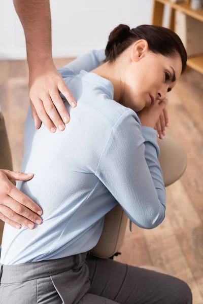Vista de cerca del masajista que masajea lastimando la espalda de una mujer de negocios sentada en una silla de masaje sobre un fondo borroso - foto de stock