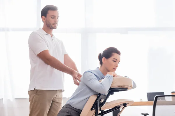 Masajista enfocado masajeando la espalda de morena de mujer de negocios sentada en silla de masaje sobre fondo borroso - foto de stock