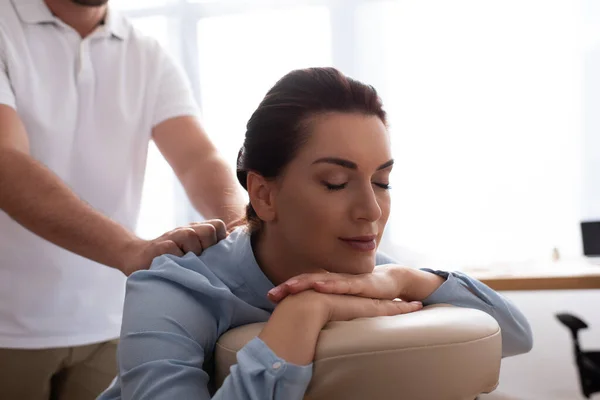 Massagista massagista massageando de volta de cliente feminino positivo com olhos fechados em fundo turvo — Fotografia de Stock