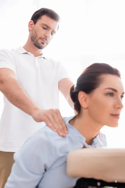 Сосредоточенный массажист делает массаж плеч позитивной клиентки на размытом переднем плане — стоковое фото