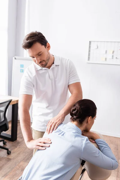 Sorridente massaggiatrice massaggiatrice che massaggia la schiena di una donna d'affari seduta sulla sedia da massaggio in ufficio — Foto stock