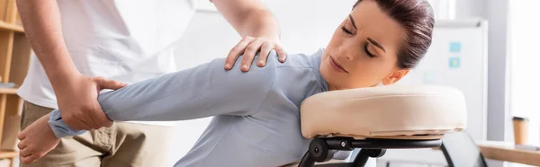 Masseur massaging painful arm of brunette businesswoman sitting on massage chair with blurred office on background, banner — Stock Photo