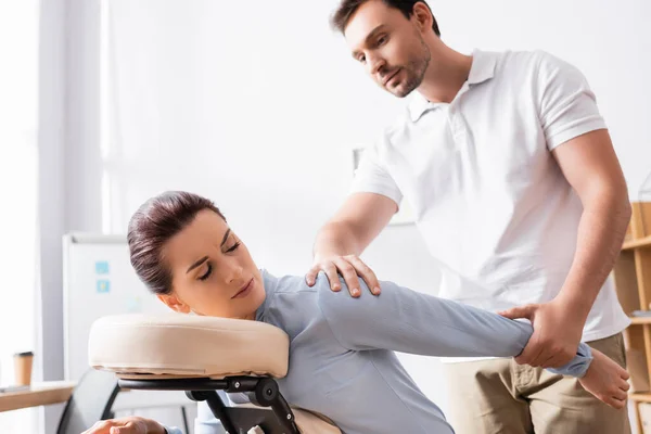 Masajista masajista brazo herido de cliente femenino sentado en silla de masaje con oficina borrosa en el fondo - foto de stock