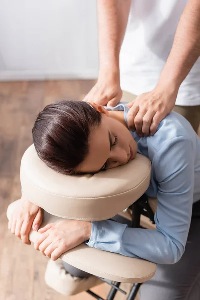 Masseur com as mãos estendidas massageando ombros de empresária morena sentada na cadeira de massagem — Fotografia de Stock