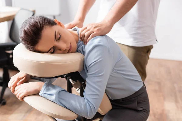 Massothérapeute massant les épaules d'une femme aux yeux fermés, assise sur une chaise de massage avec bureau sur fond flou — Photo de stock