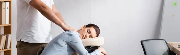 Vista lateral del masajista masajeando hombros de mujer con los ojos cerrados sentado en silla de masaje en la oficina, pancarta - foto de stock