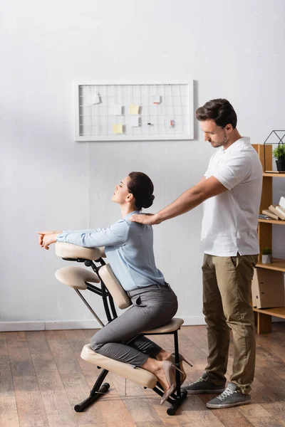 Вид сбоку массажиста с протянутыми руками, делающего массаж плеч деловой женщины, сидящей на массажном кресле в офисе — стоковое фото