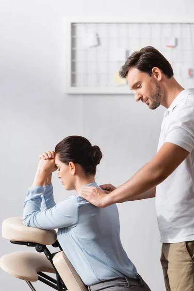 Massaggiatore sorridente che fa massaggio seduto di spalle di donna d'affari in ufficio su sfondo sfocato — Foto stock