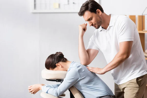 Masseur souriant massant femme d'affaires retour avec coude au bureau sur fond flou — Photo de stock