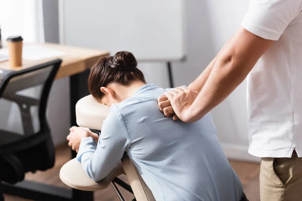 Massagista massageando mulher morena de volta sentado na cadeira de massagem com escritório borrado no fundo — Fotografia de Stock