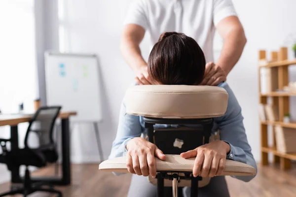 Vista frontal do massagista fazendo massagem sentada de ombros para mulher de negócios com escritório embaçado no fundo — Fotografia de Stock