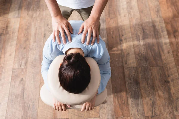 Верхний вид массажера, делающего сидячий массаж плеч брюнетке на текстурированном фоне — стоковое фото