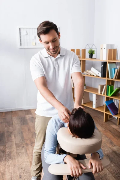 Massagista sorridente fazendo massagem sentada no pescoço para empresária morena no escritório — Fotografia de Stock