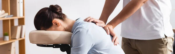 Side view of masseur massaging back of brunette woman sitting on massage chair on blurred background, banner — Stock Photo