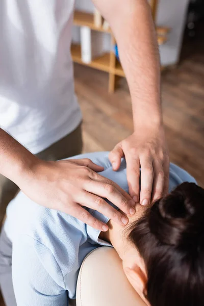 Vista recortada de masajista haciendo masaje sentado de cuello para mujer morena con oficina borrosa sobre fondo - foto de stock