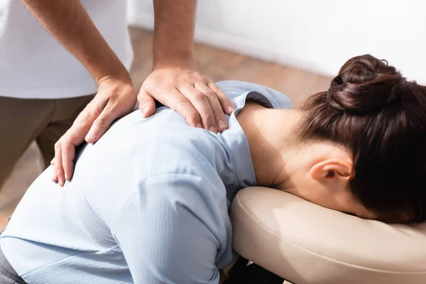 Vue rapprochée du massothérapeute faisant un massage assis du dos pour femme brune sur fond flou — Photo de stock