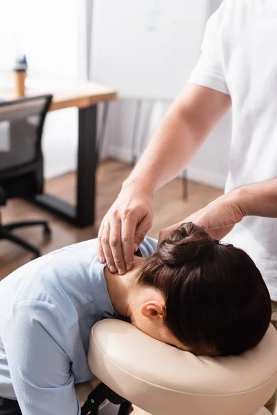 Массажный терапевт массирует шею брюнетки бизнес-леди, сидящей на массажном кресле на размытом фоне — стоковое фото