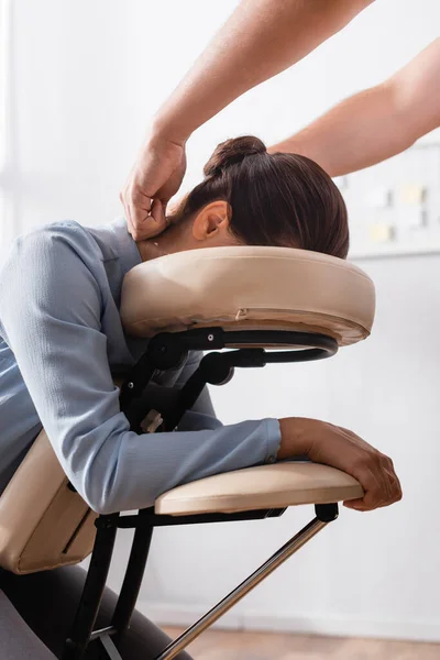 Masajista haciendo masaje sentado de cuello para mujer de negocios morena sobre fondo borroso - foto de stock