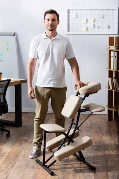 Vista frontal del masajista sonriente mirando a la cámara, mientras está de pie cerca de la silla de masaje en la oficina - foto de stock