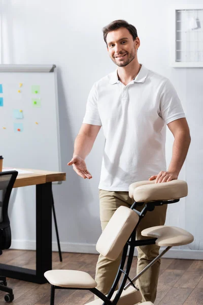 Vue de face du masseur joyeux debout et pointant avec la main sur la chaise de massage dans le bureau — Photo de stock