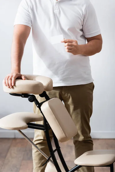 Cropped view of masseur standing near massage chair and holding blank business card — Stock Photo