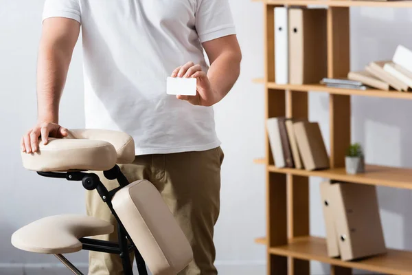 Обрізаний вид масажиста, що стоїть біля масажного крісла і тримає порожню візитку в офісі на розмитому фоні — стокове фото