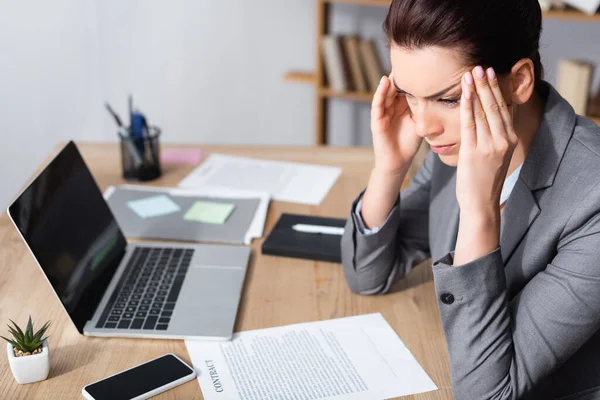 Businesswoman with migraine looking away while sitting near contracts and laptop on table on blurred background — Stock Photo