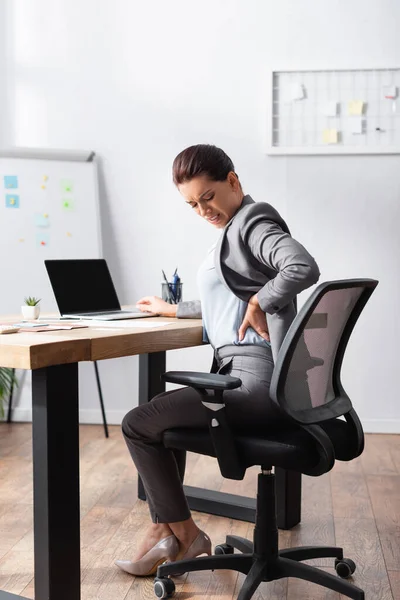 Mujer de negocios agotada con dolor de espalda cogida de la mano en la cadera, mientras se sienta en la silla de la oficina en la oficina - foto de stock