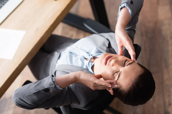 Überkopfansicht einer erschöpften Geschäftsfrau mit Migräne, die auf einem Bürostuhl auf verschwommenem Hintergrund sitzt — Stockfoto