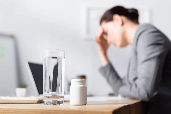 Glas Wasser und Medikamente auf dem Tisch mit verschwommener Geschäftsfrau mit Migräne im Hintergrund — Stockfoto