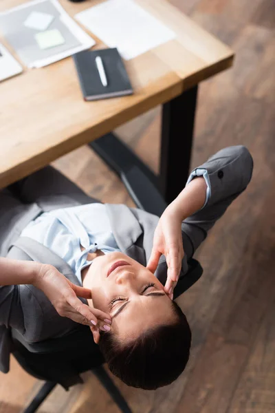 Vista aérea de la trabajadora de oficina con dolor de cabeza sentada en la silla de oficina sobre fondo borroso - foto de stock