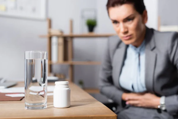 Glas Wasser und Medikamente auf dem Schreibtisch mit verschwommener Geschäftsfrau mit Bauchschmerzen im Hintergrund — Stockfoto