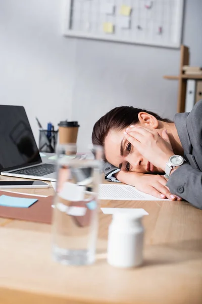Mujer de negocios agotada con dolor de cabeza apoyada en el escritorio con contrato y portátil en primer plano borroso - foto de stock