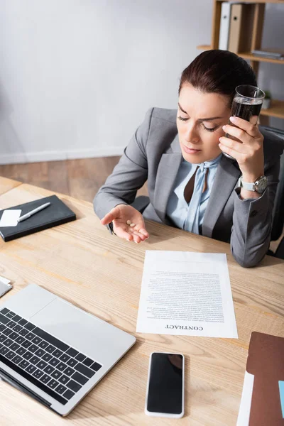 Müde Geschäftsfrau hält ein Glas Wasser in der Hand und betrachtet Schmerzmittel, während sie am Arbeitsplatz im Büro sitzt — Stock Photo