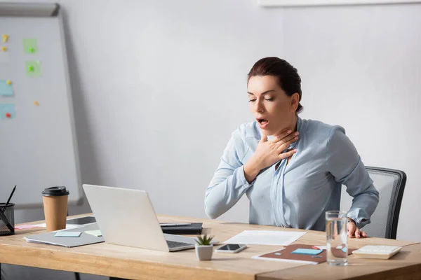 Brunette businesswoman with hand on neck coughing while sitting at workplace — Stock Photo