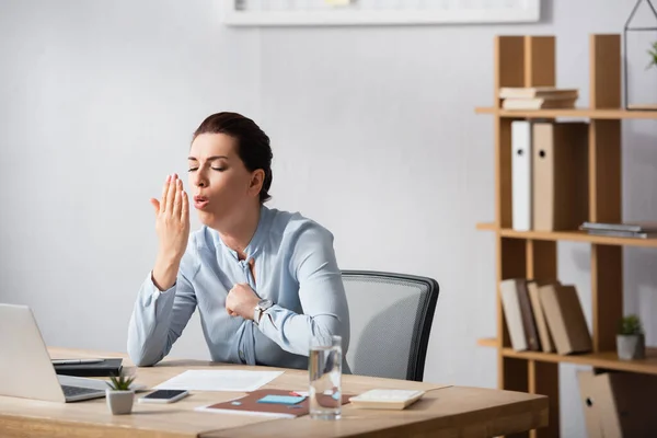Brunette businesswoman with fist on chest coughing on palm while sitting at workplace in office — Stock Photo