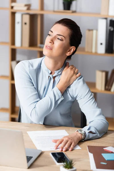 Mujer de negocios cansada con el hombro dolorido mirando hacia otro lado, mientras está sentada en el lugar de trabajo sobre un fondo borroso - foto de stock