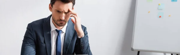 Tired businessman with headache, holding hand near temple and looking away with blurred flipchart on background, banner — Stock Photo