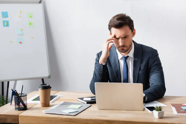 Homme d'affaires avec migraine regardant ordinateur portable tout en étant assis sur le lieu de travail près de tableau à feuilles mobiles — Photo de stock