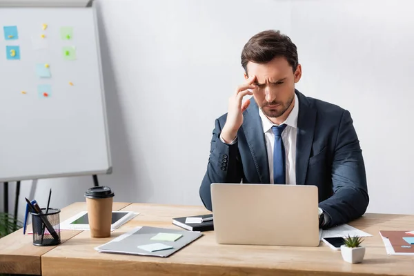 Uomo d'affari con emicrania guardando computer portatile mentre seduto sul posto di lavoro vicino flipchart — Foto stock