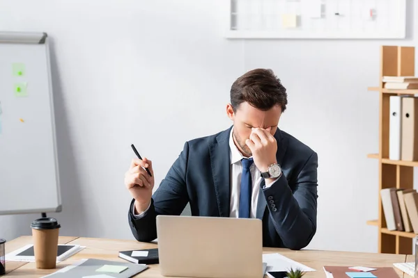 Hombre de negocios agotado con dolor de cabeza, cogido de la mano cerca de los ojos, sentado en el lugar de trabajo sobre fondo borroso - foto de stock